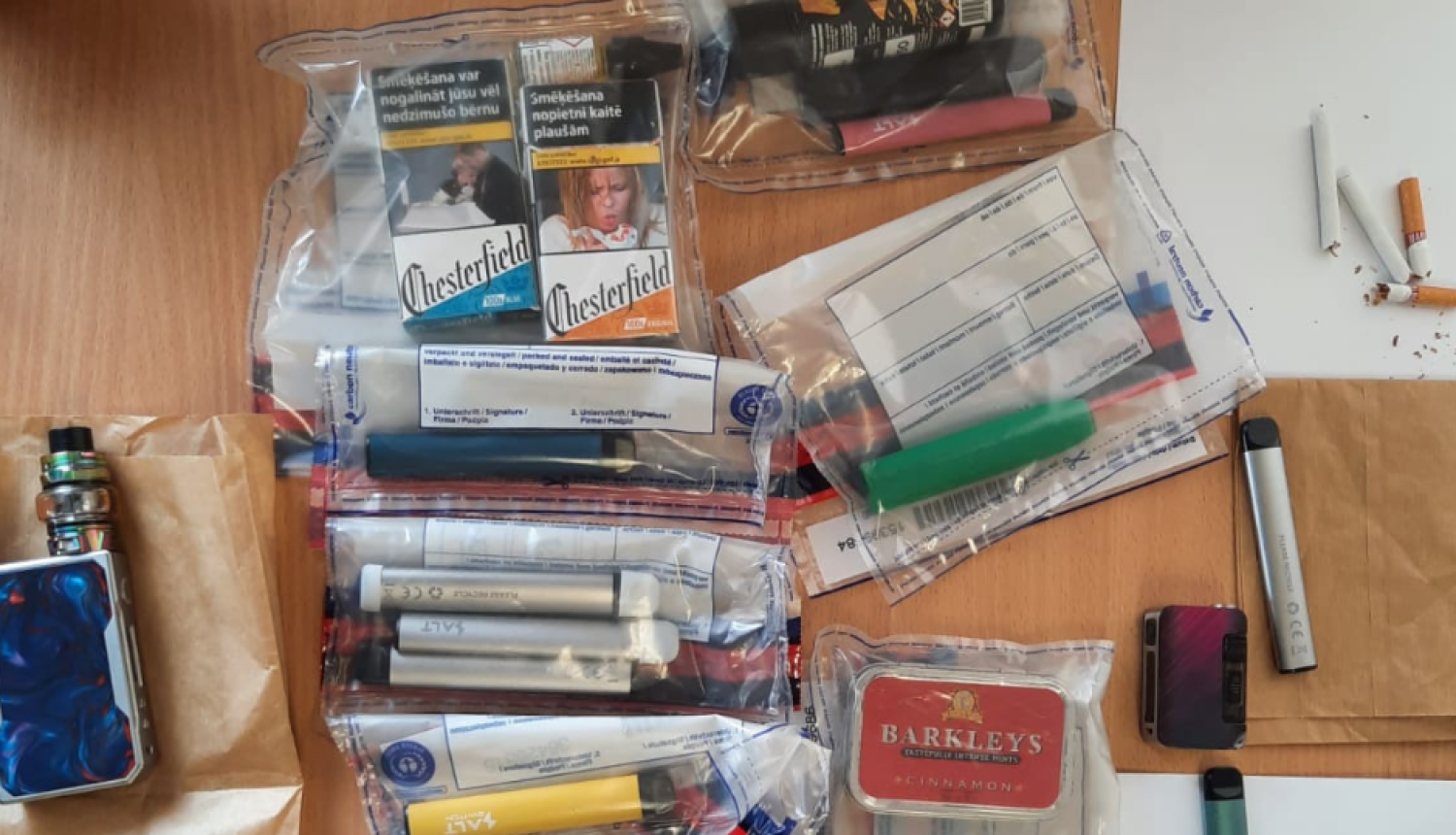 Uz galda vairākos caurspīdīgos plastmasas maisiņos iepakotas elektroniskās cigaretes, redzamas arī dažas cigarešu paciņas un uz baltas papīra lapas augšējā labā stūrī trīs atsevišķas cigaretes