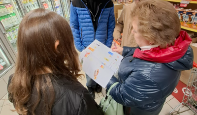 Četri cilvēki veikalā, sievietei rokās papīra lapas, uz kurām attēloti pārtikas produkti