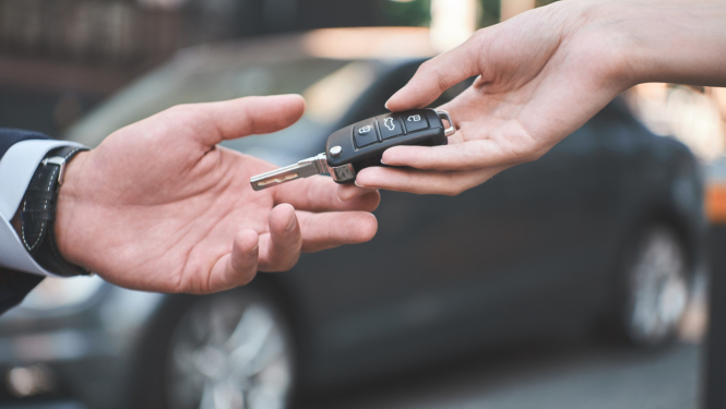 Fomā melna automašīna, priekšplānā divu cilvēku rokas, kas pasniedz automašīnas atslēgas