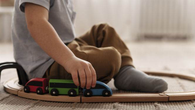 Zēns sēžu uz grīdas, spēlējas ar rotaļu vilcieniņu. Apkārt zēnam iet vilcieniņa sliedes.