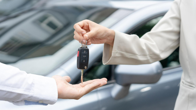 No rokas rokā tiek nodota automašīnas atslēga, fonā pelēcīgs auto