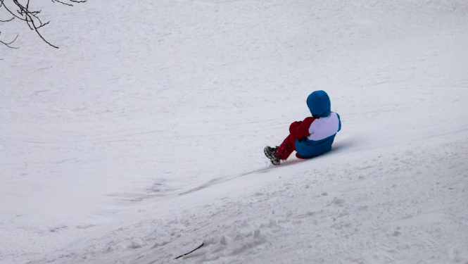 Kalns ar sniegu, no kura uz ragaviņām šļūc bērns. Bērns redzams no mugurpuses.