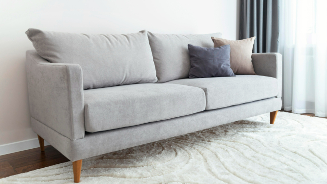 Pelēks dīvāns, uz tā divi spilveni tumši pelēkā un brūnganā tonī, aizmugurē pelēki aizkari