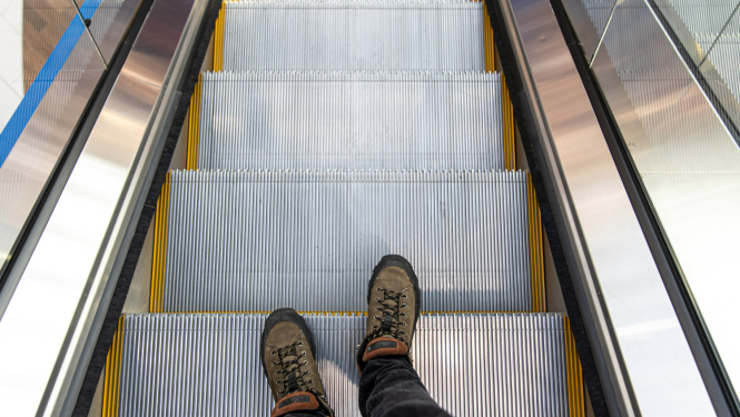 Eskalators, uz viena pakāpiena redzamas vīrieša kājas brūnās kurpēs