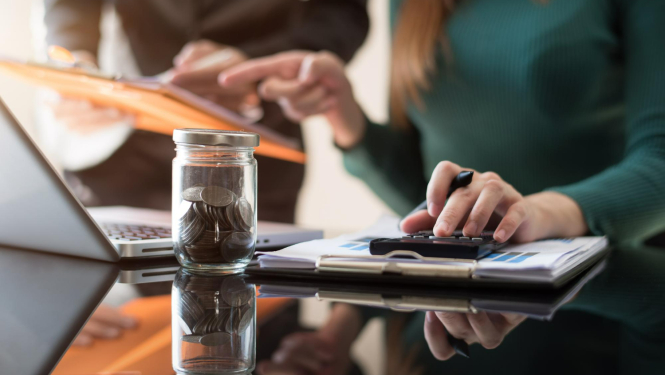 Divi cilvēki pie datora, notiek konsultācija, priekšplānā trauks ar monētām