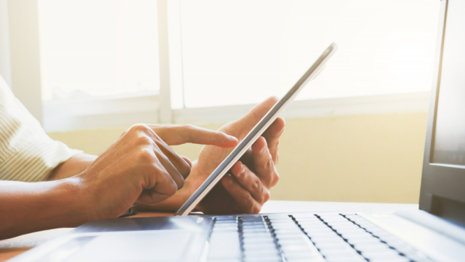 Planšete sievietes rokās, blakus atvērts dators