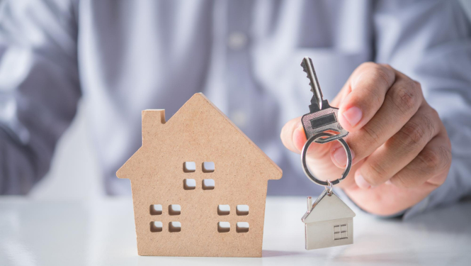 Priekšplānā mājas stiluets no kartona, tai blakus vīrieša roka, kas tur atslēgu ar piekariņu mājās veidolā