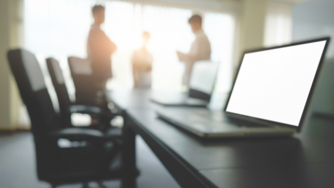 Sanāksmes telpa, uz galda priekšplānā atvērts dators, fonā stāv 3 cilvēki