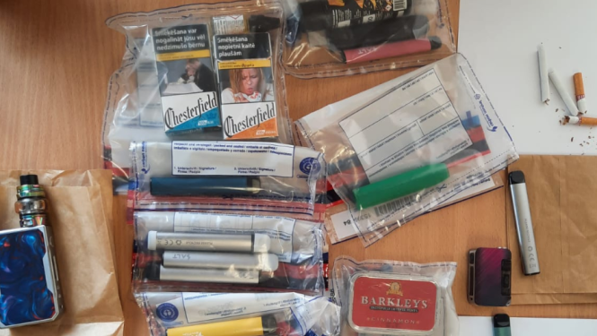 Uz galda vairākos caurspīdīgos plastmasas maisiņos iepakotas elektroniskās cigaretes, redzamas arī dažas cigarešu paciņas un uz baltas papīra lapas augšējā labā stūrī trīs atsevišķas cigaretes
