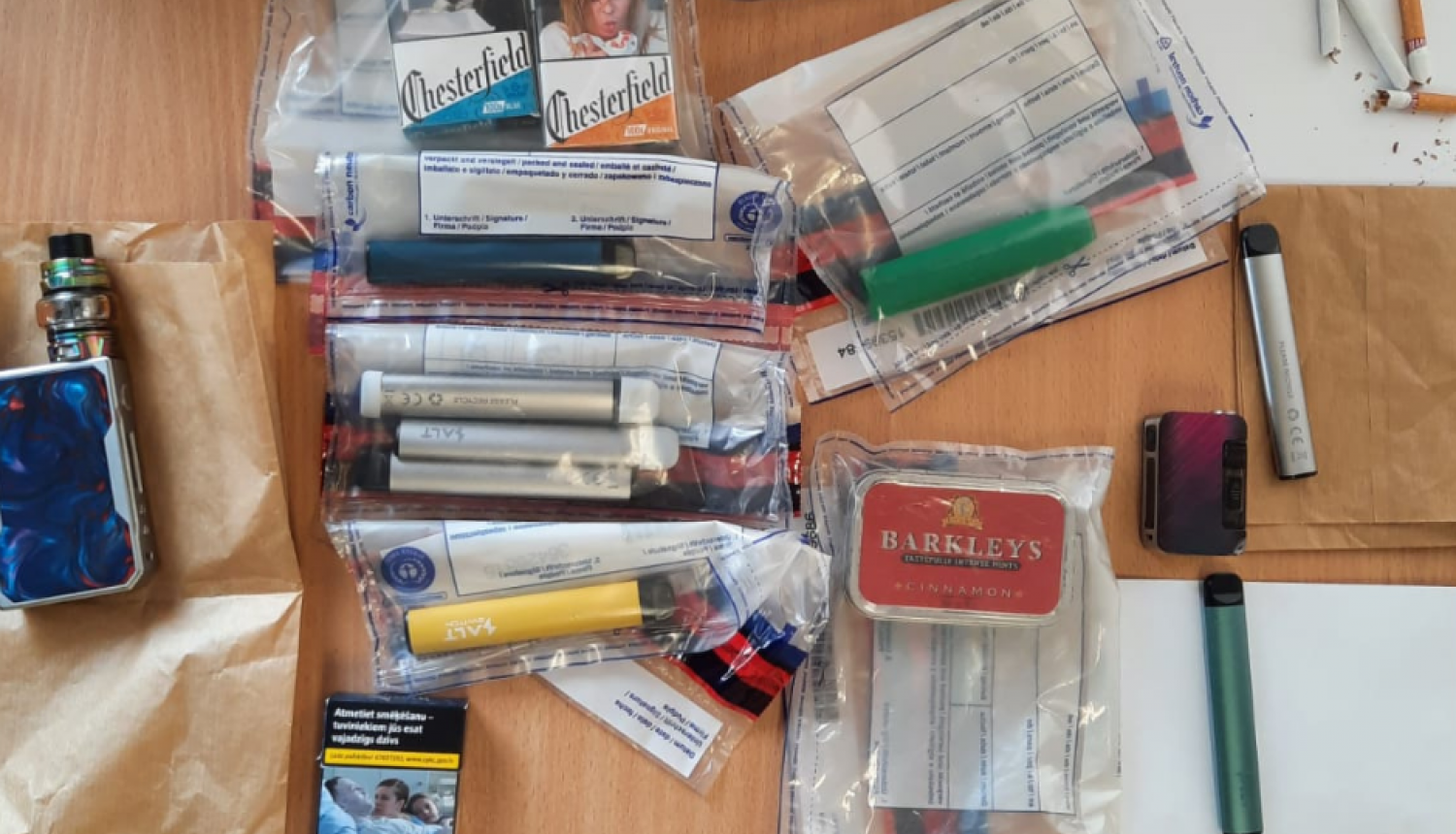 Uz galda vairākos caurspīdīgos plastmasas maisiņos iepakotas elektroniskās cigaretes, redzamas arī dažas cigarešu paciņas un uz baltas papīra lapas augšējā labā stūrī trīs atsevišķas cigaretes